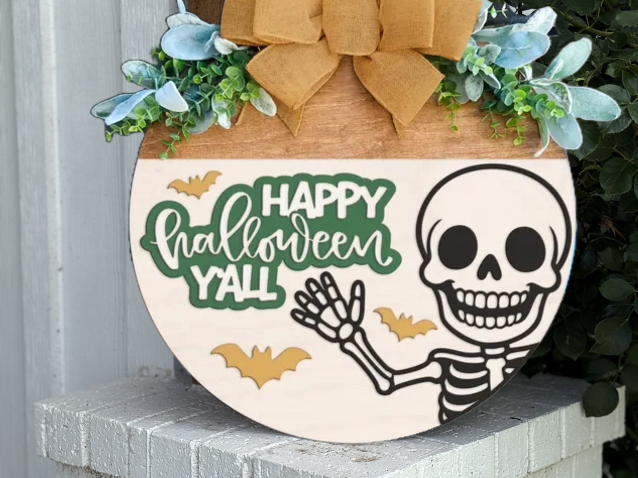 A round wooden sign decorated with greenery, a tan bow, and featuring a smiling skeleton, yellow bats, and the words Happy Halloween Yall in playful green and white letters.