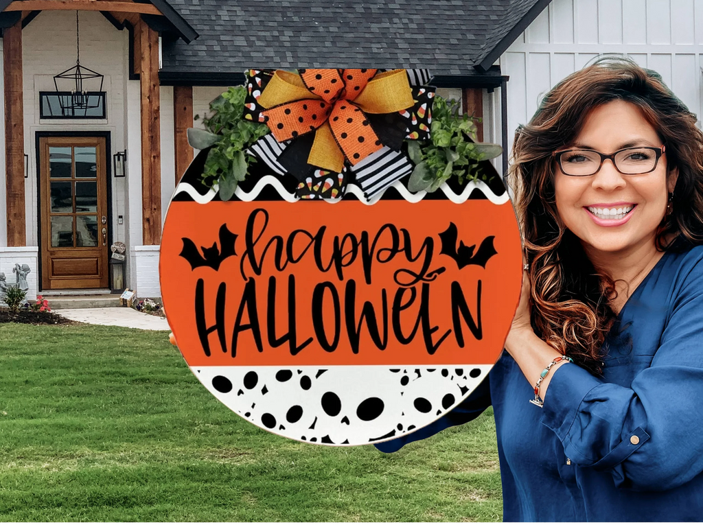 A smiling woman with wavy brown hair and glasses stands outside a house next to a festive round sign that reads Happy Halloween with bat illustrations and a large orange and black bow on top.