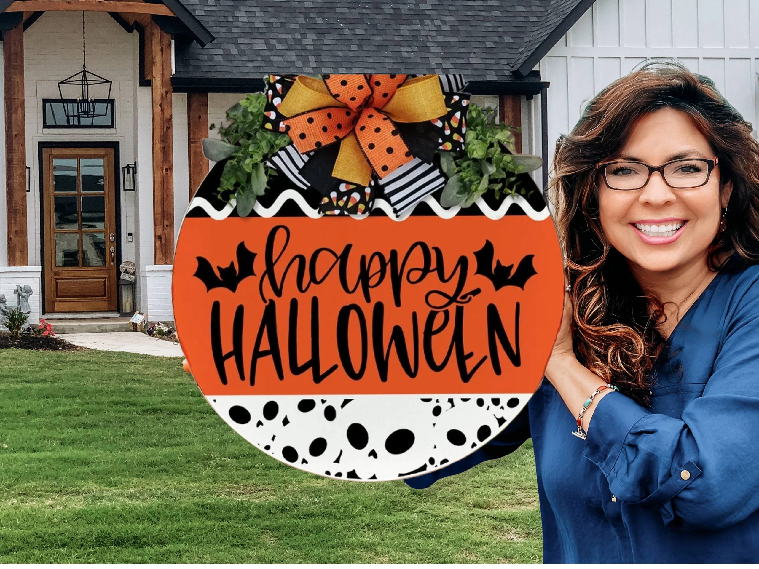 A smiling woman with wavy brown hair and glasses stands outside a house next to a festive round sign that reads Happy Halloween with bat illustrations and a large orange and black bow on top.