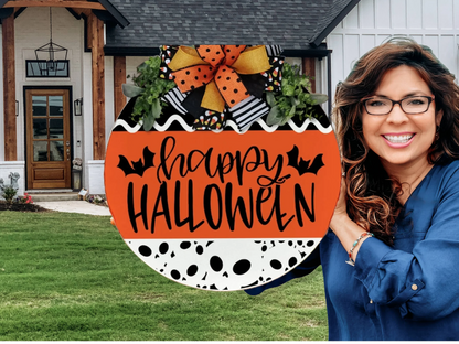 A smiling woman with wavy brown hair and glasses stands outside a house next to a festive round sign that reads Happy Halloween with bat illustrations and a large orange and black bow on top.