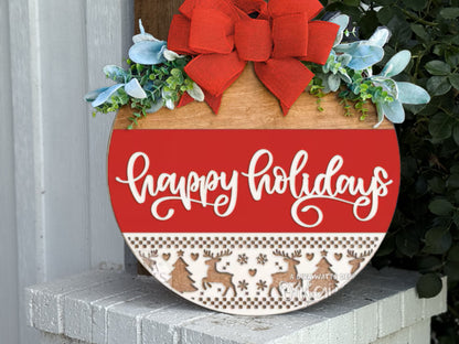 A round wooden holiday sign with a red bow, greenery, and the words happy holidays in white script. The bottom features a pattern of reindeer, trees, and snowflakes. The sign rests on a white brick surface.
