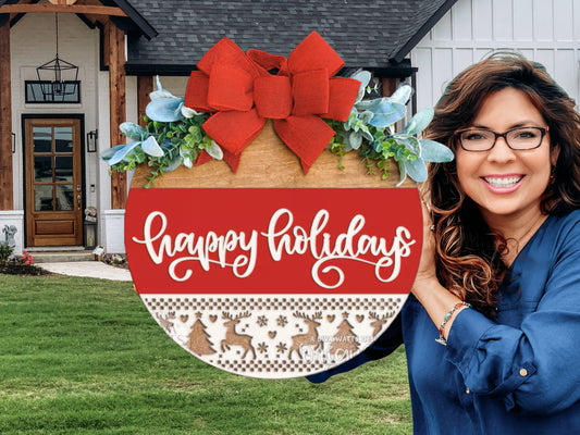 A smiling woman holds a round holiday sign with a red bow, greenery, and happy holidays written in script. She stands in front of a modern house with a wooden door and white siding.