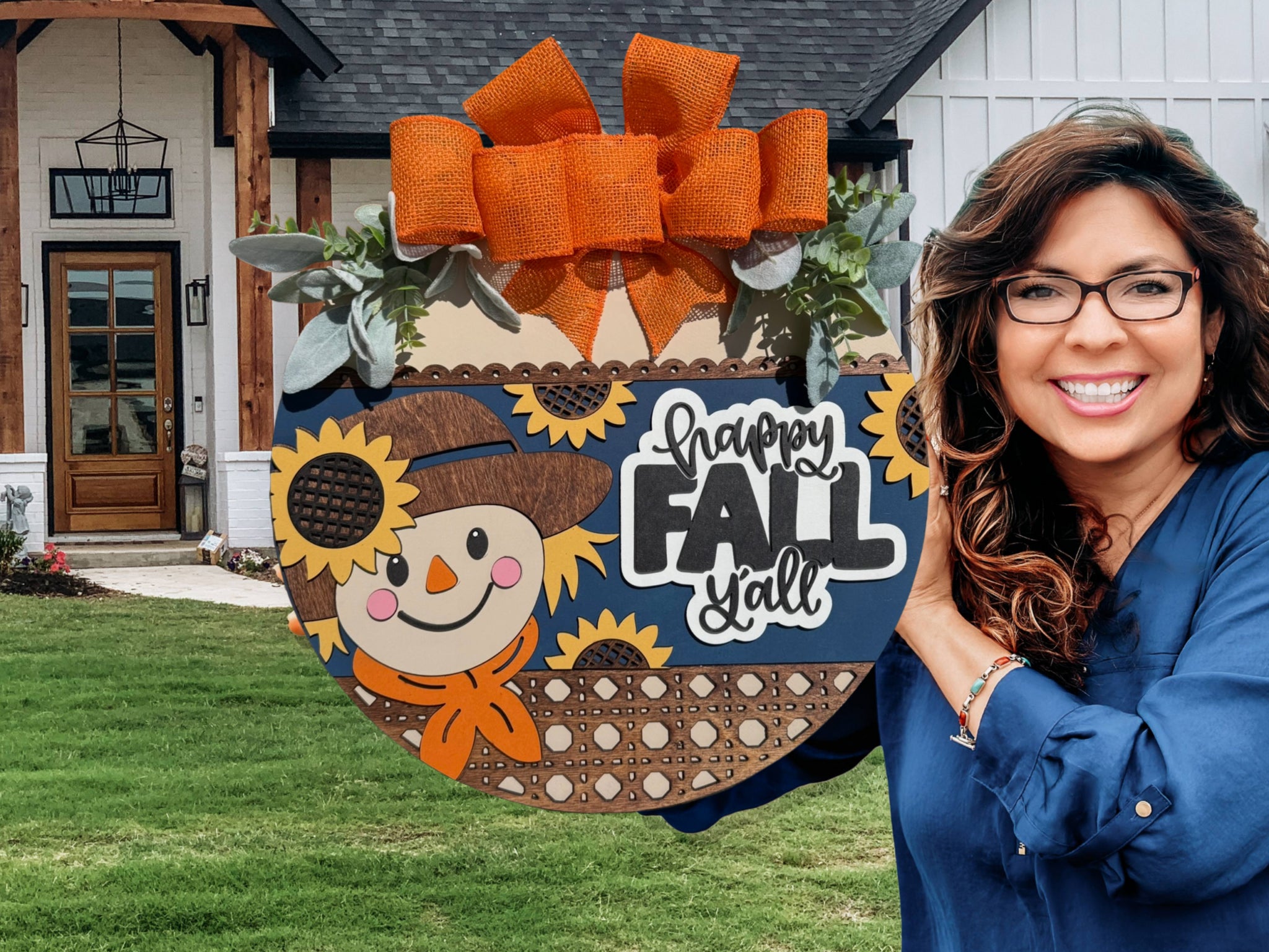 A smiling woman with long brown hair and glasses holds a round wooden sign decorated with a scarecrow, sunflowers, orange bow, and the words happy FALL yall in front of a house with a green lawn.
