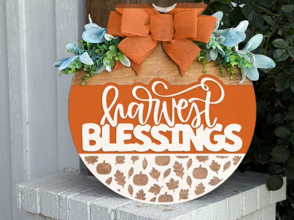 A round wooden sign decorated with greenery, blue leaves, and an orange bow. It reads harvest BLESSINGS in white script, with a bottom section displaying a pattern of pumpkins and leaves. The sign rests on a brick ledge.
