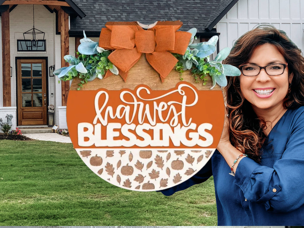 A round wooden sign decorated with greenery, blue leaves, and an orange bow. It reads harvest BLESSINGS in white script, with a bottom section displaying a pattern of pumpkins and leaves. The sign rests on a brick ledge.