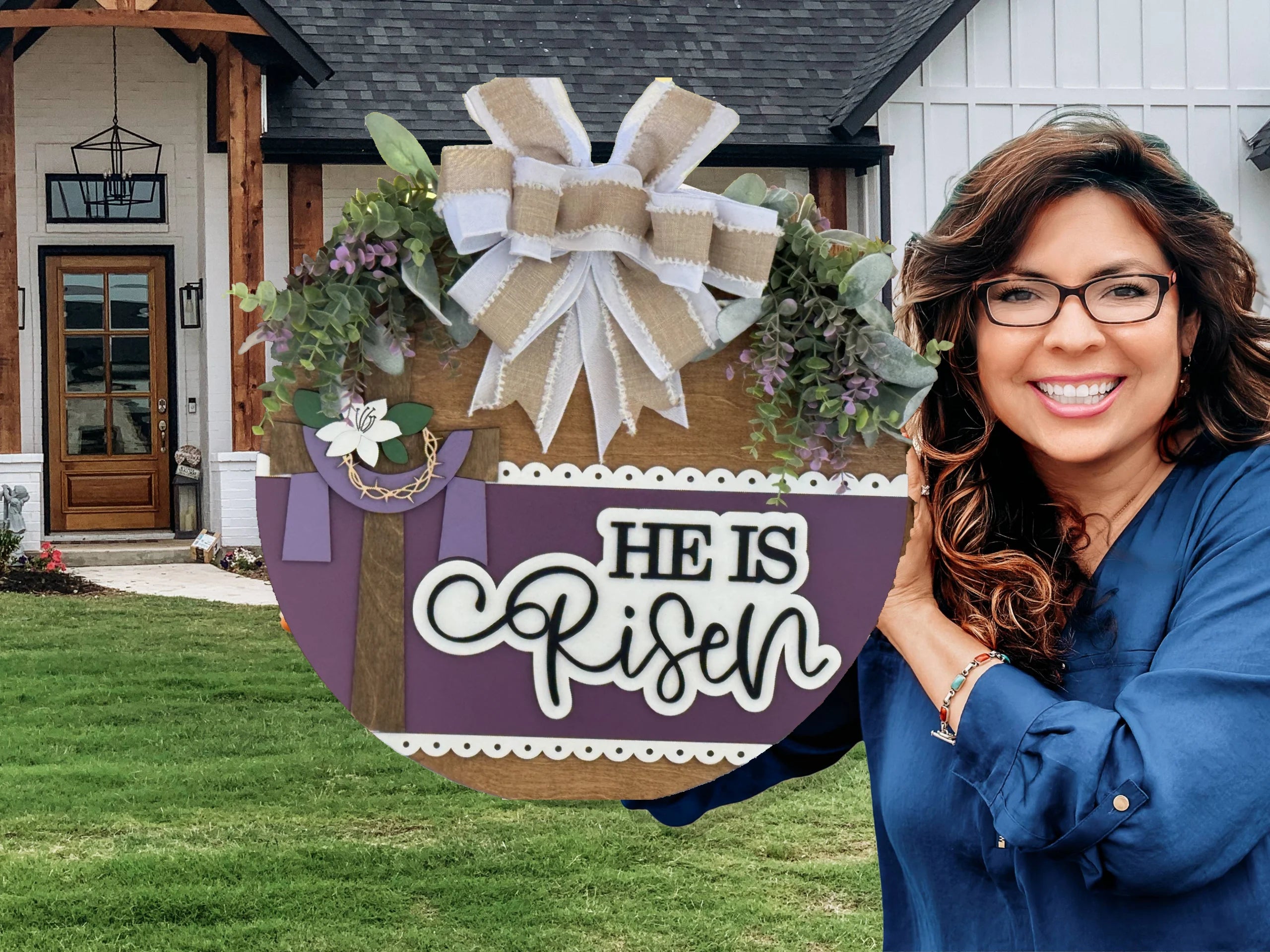 A smiling woman with long brown hair and glasses stands outside a house, holding a round sign decorated with greenery, a cross, and a large bow. The sign reads “HE IS Risen” in bold and script white letters on a purple background.