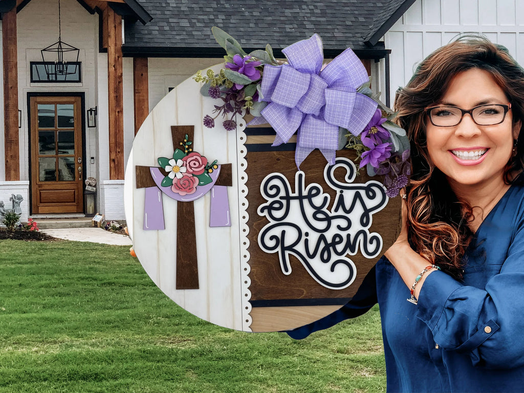 A smiling woman with wavy brown hair and glasses holds a circular wooden sign decorated with a cross, flowers, purple bow, and the words He is Risen. She stands outside in front of a modern house with a wooden door and flowerbeds.