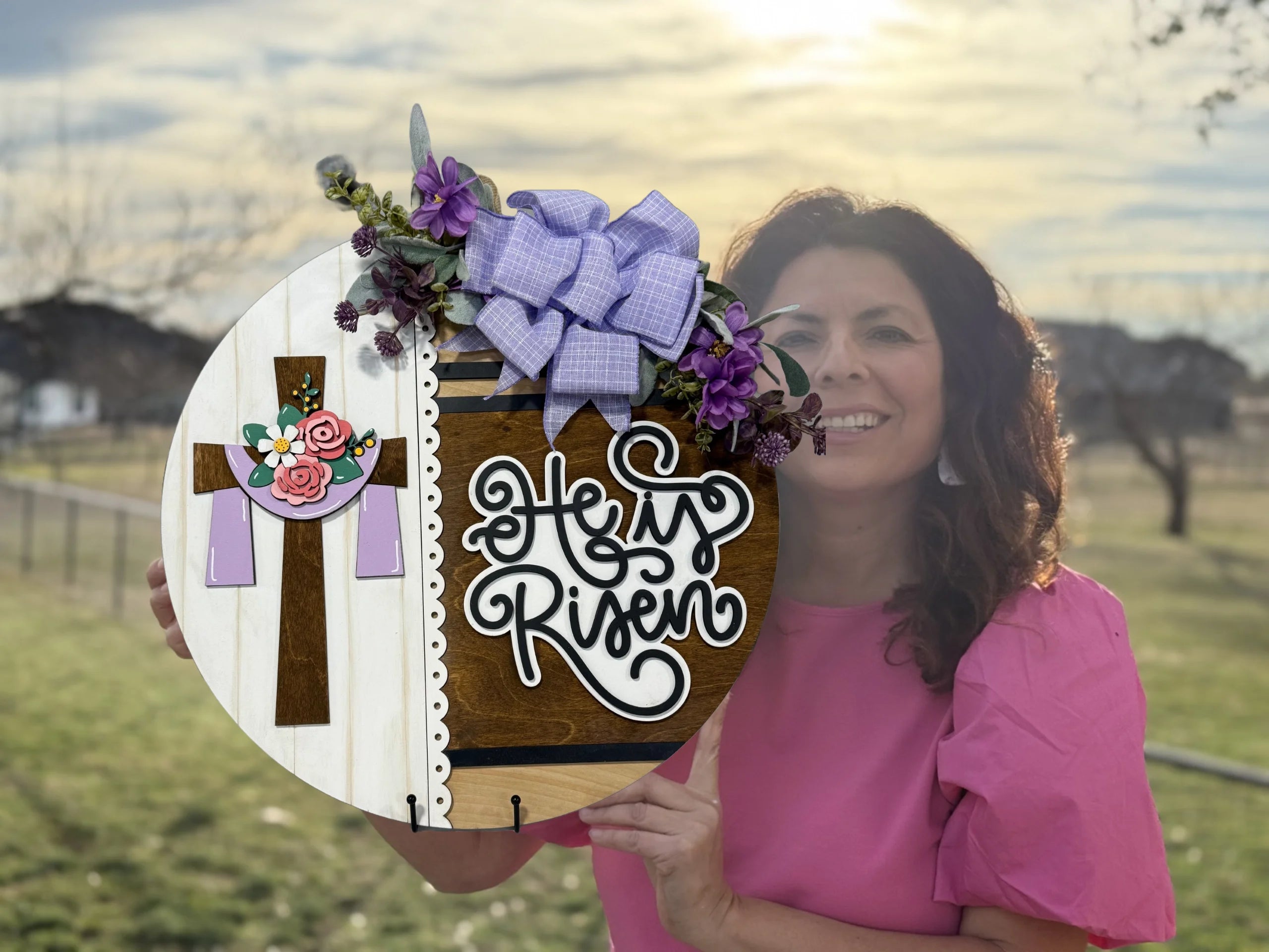 A woman in a pink blouse holds a round wooden Easter sign featuring a cross with flowers and the text He is Risen. The sign is decorated with a purple bow and floral accents. The background showcases a grass field and a softly lit sky with trees in the distance.