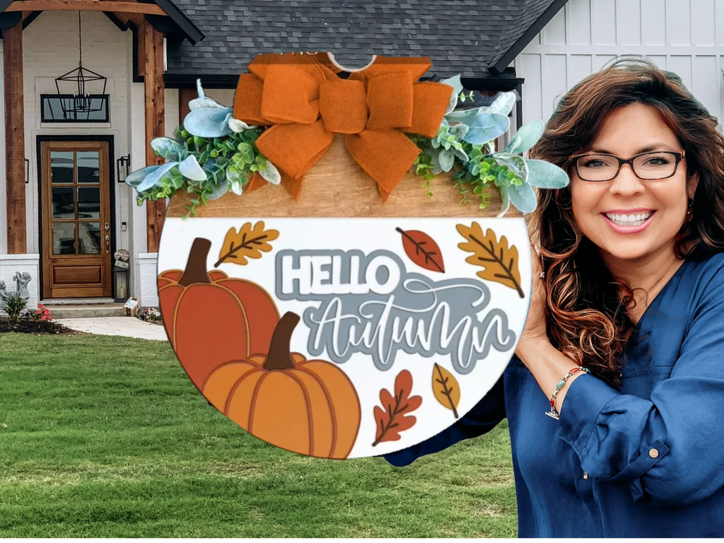 A woman with long brown hair and glasses, smiling, holds a round autumn-themed sign that reads Hello Autumn with pumpkins and leaves. She stands in front of a modern house with a black door and white siding.