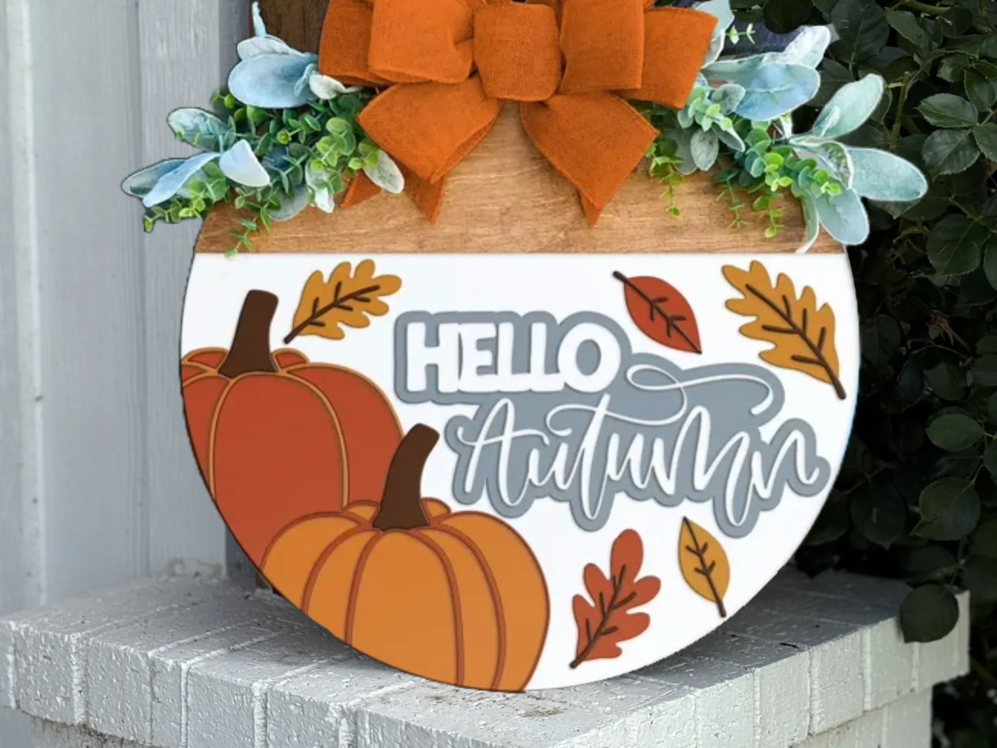 A round wooden sign decorated with a large orange bow, leaves, greenery, two orange pumpkins, and the words “Hello Autumn” in white and gray, hanging outside on a white brick ledge.