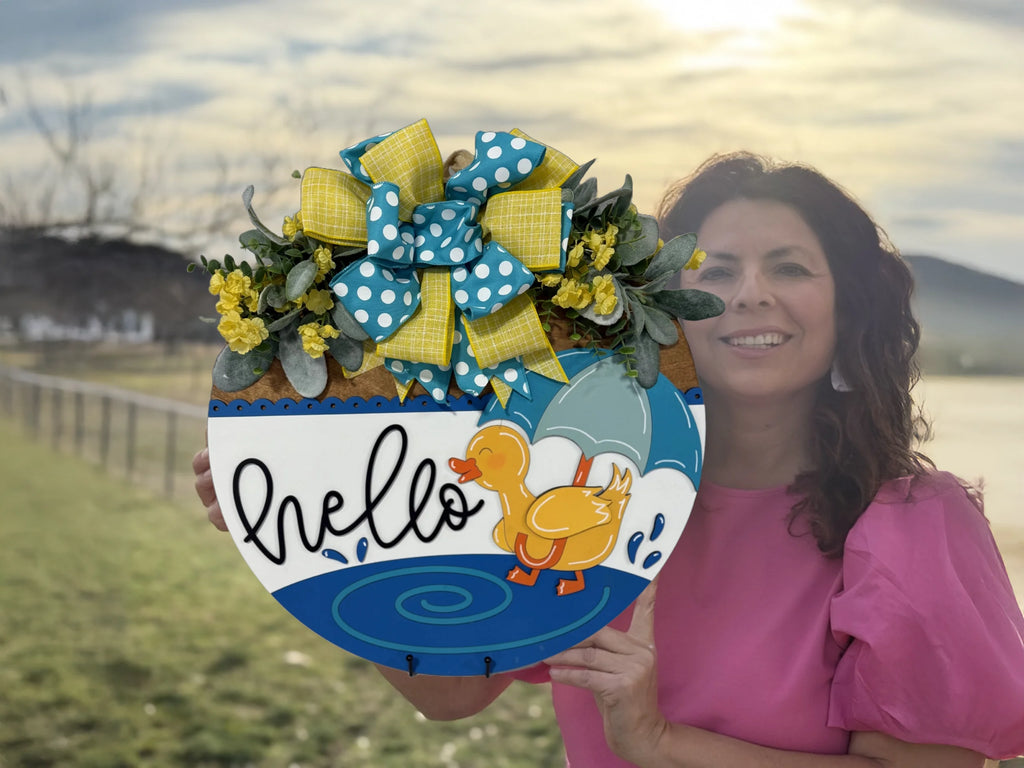 A woman in a pink shirt holds a circular, spring-themed welcome sign against a sunny, rural backdrop. The sign features a decorated bow, a duck with an umbrella, and the word hello in artistic script. The sky is bright with scattered clouds and a hint of sun.