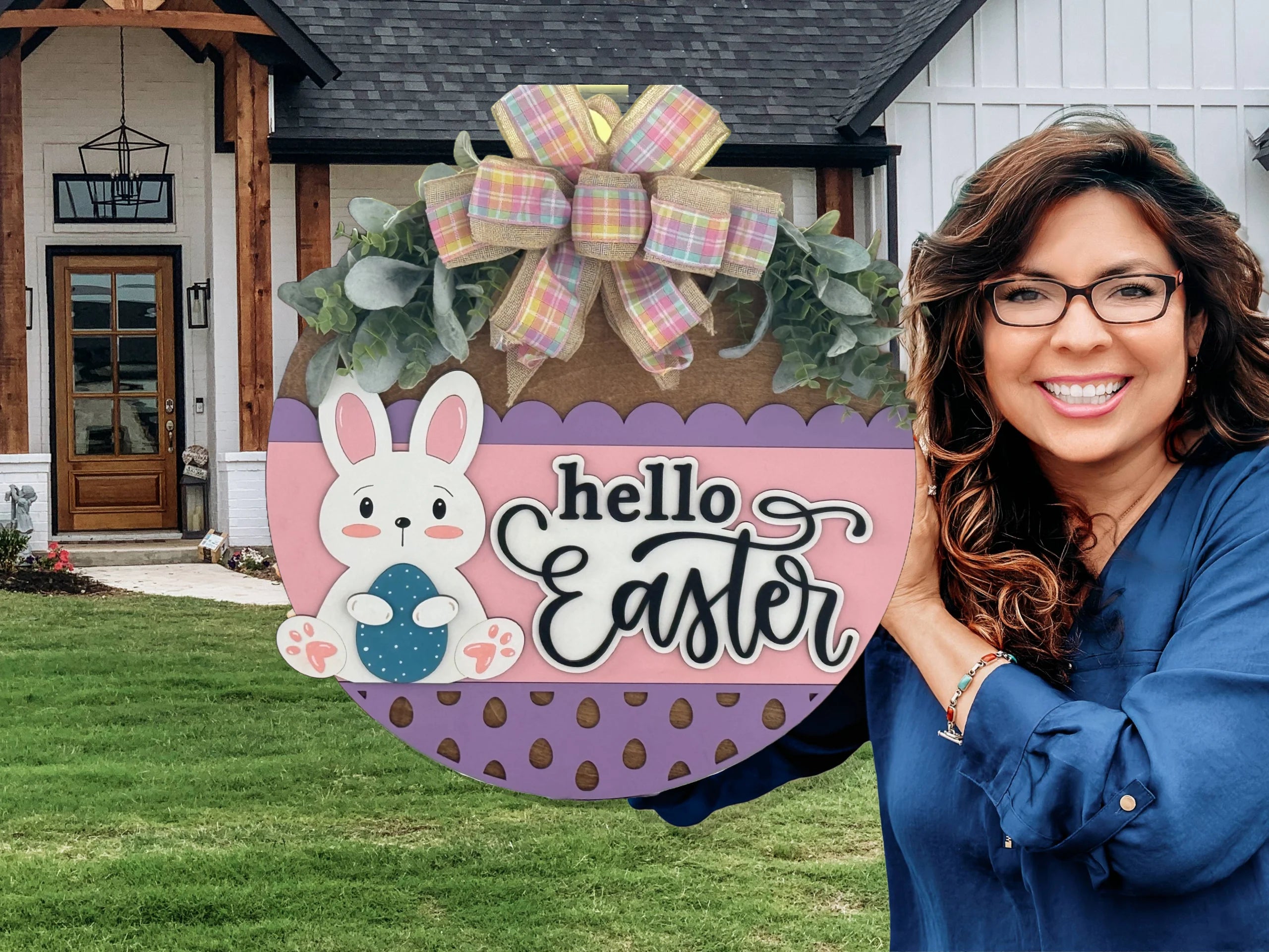 A smiling woman with wavy brown hair and glasses holds a round, pastel-colored Hello Easter sign decorated with a bunny, polka dots, greenery, and a plaid bow, standing outside a modern house with a porch and green lawn.