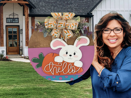 A smiling woman with wavy brown hair holds a round decorative sign. The sign features a cartoon bunny with a carrot, a “hello” script, and a large carrot-themed bow. She stands in front of a modern house with a wooden porch and green lawn.