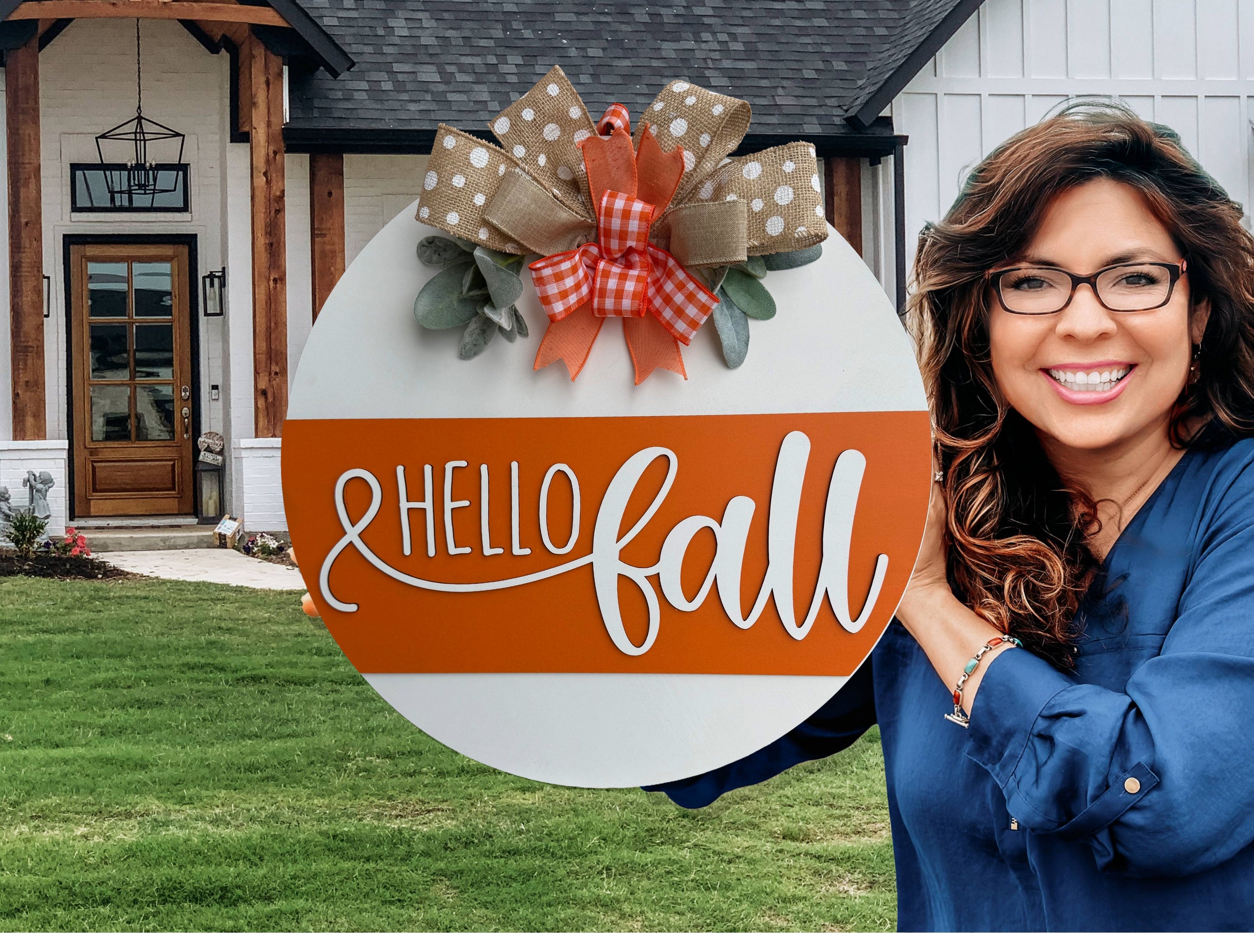 A round sign with Hello Fall in white script on an orange stripe, decorated with polka dot and checkered bows and faux greenery, is displayed on a white brick ledge near a garden.