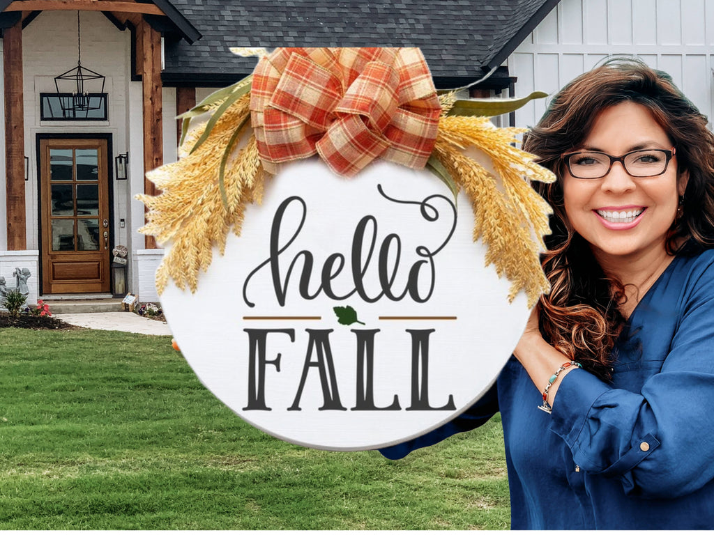 A smiling woman with long brown hair and glasses holds a round sign decorated with a plaid bow and wheat that reads hello FALL in front of a modern farmhouse-style home.