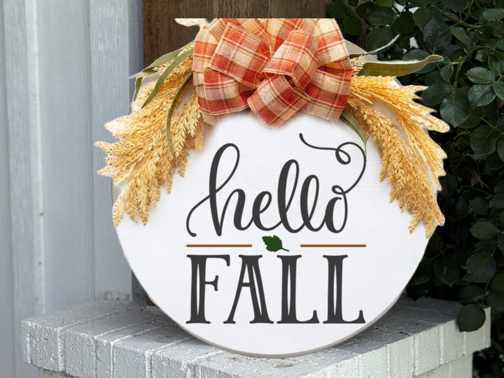 A round sign with hello FALL written on it, decorated with a large orange plaid bow and wheat stalks, is displayed on a white brick ledge.