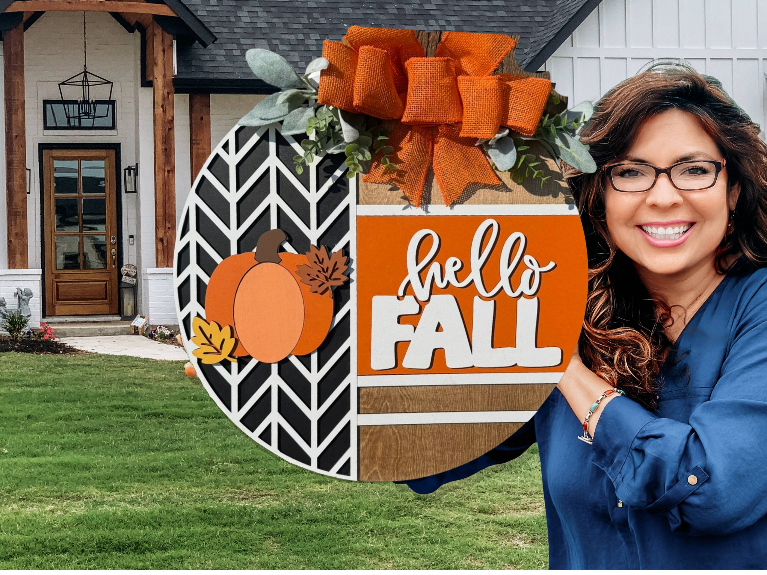 A smiling woman with wavy hair holds a round sign decorated with pumpkins, leaves, and an orange bow that reads hello FALL in front of a modern house with a green lawn.