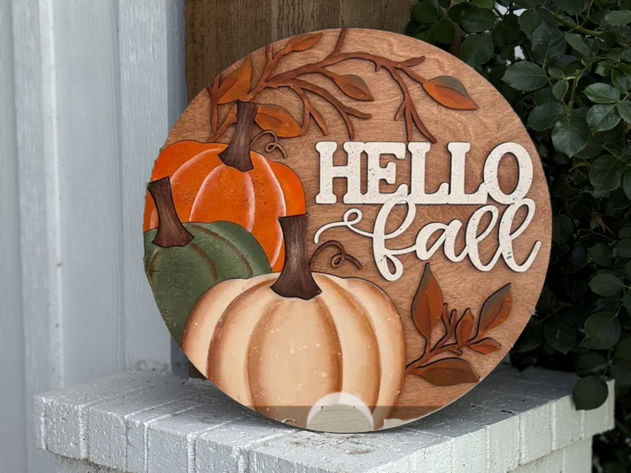 A round wooden sign with HELLO fall written in white, decorated with painted pumpkins in orange, green, and cream, and autumn leaves, propped on a white brick ledge near green foliage.