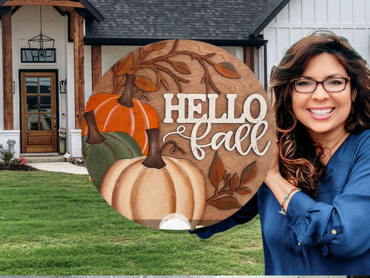 A smiling woman in glasses holds a round wooden sign with pumpkins and leaves that says HELLO fall, standing outside a modern house with a dark roof and wooden accents.