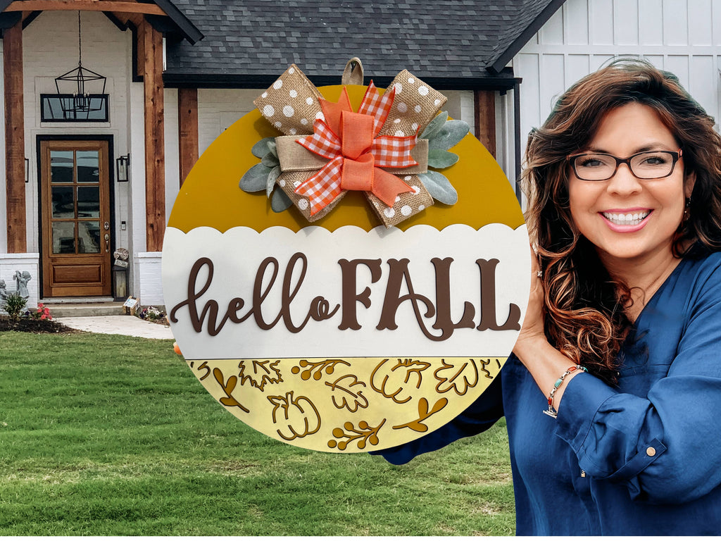 A round yellow and white sign with hello FALL written on it, decorated with a polka dot and plaid bow, autumn leaves, and placed on a white brick ledge next to a wooden post and some greenery.