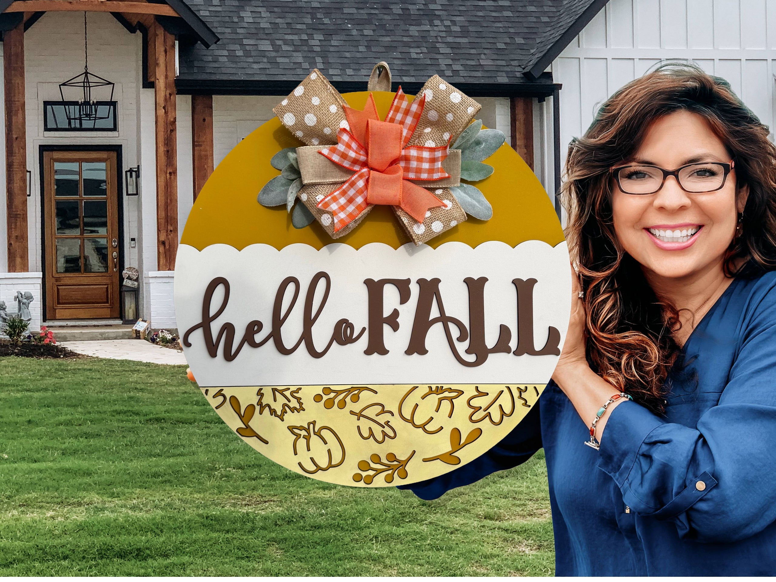 A round yellow and white sign with hello FALL written on it, decorated with a polka dot and plaid bow, autumn leaves, and placed on a white brick ledge next to a wooden post and some greenery.