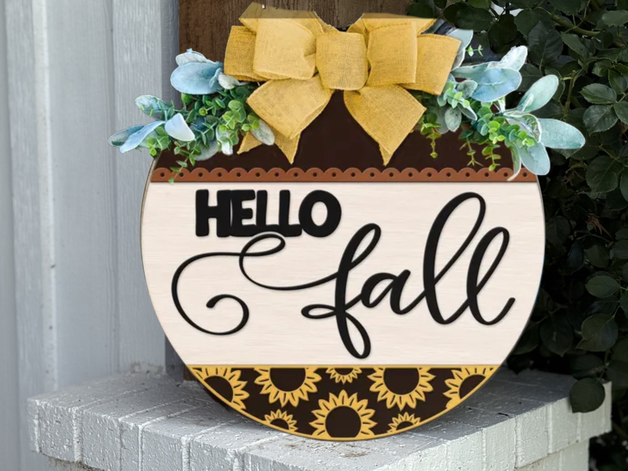 A round wooden sign decorated with sunflowers, green leaves, and a large yellow bow reads “HELLO fall” in bold and cursive letters, sitting on a white brick surface near green foliage.