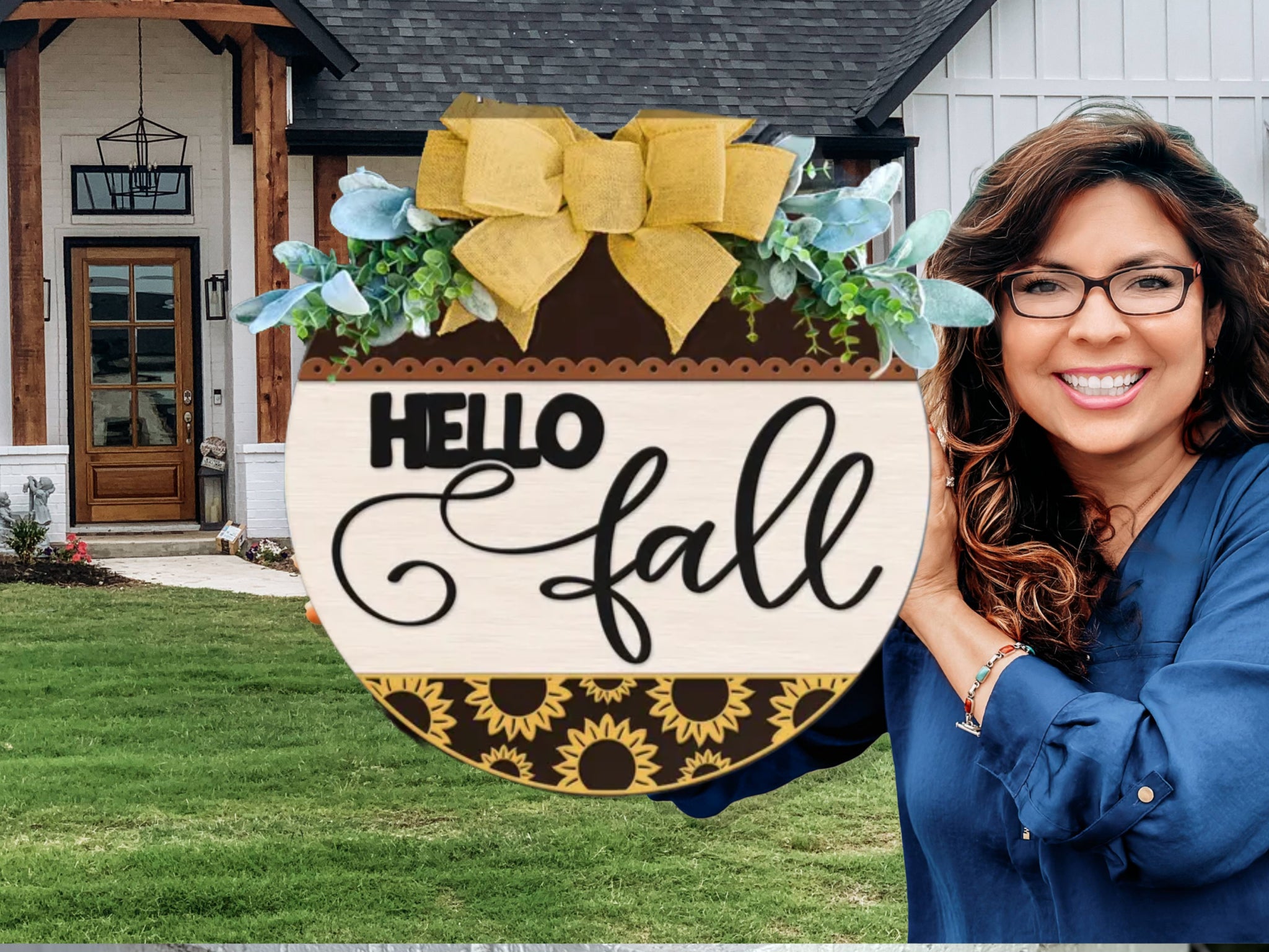 A smiling woman with long brown hair holds a round wooden sign that reads Hello Fall with decorative flowers and a yellow bow, standing in front of a modern house with a green lawn.