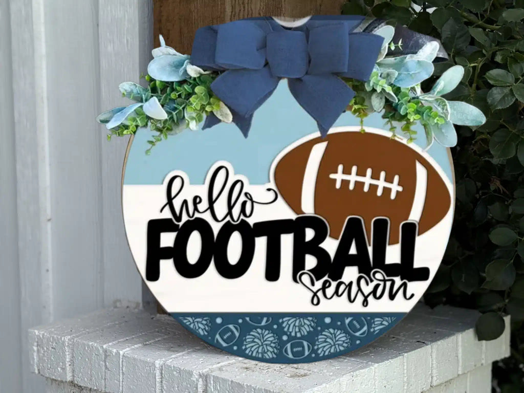 A round decorative sign with blue and white sections, greenery, and a blue bow displays the words hello FOOTBALL season next to a brown football graphic. The sign rests on a white brick surface.