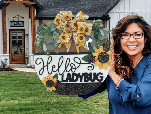 A smiling woman with wavy brown hair and glasses stands outdoors, holding a large decorative sign that reads Hello LADYBUG with painted sunflowers, green foliage, and yellow plaid bows. A modern farmhouse with wood accents is in the background.