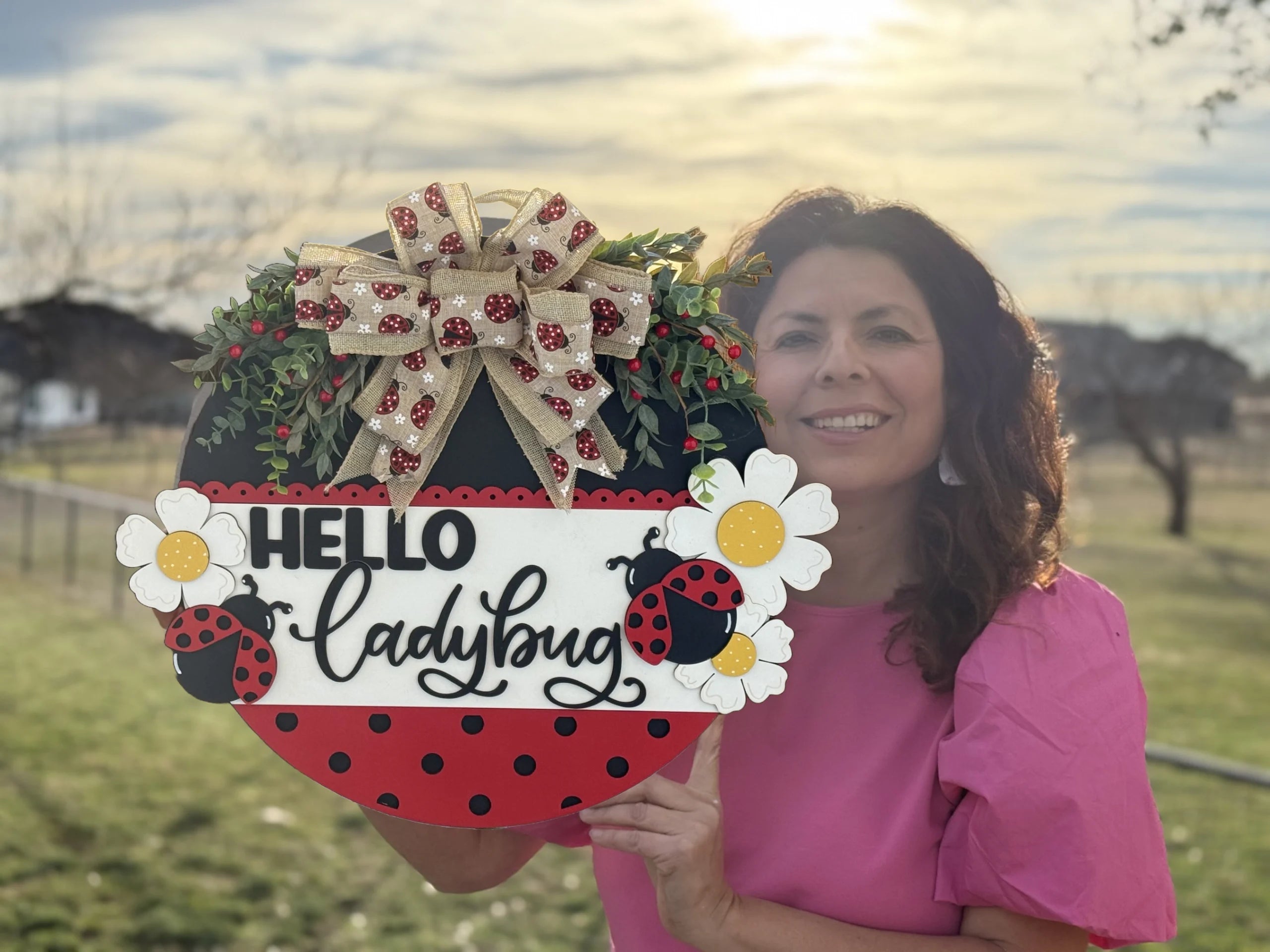 A woman in a pink shirt holds a circular sign with Hello Ladybug written on it. The sign features ladybugs, white flowers, greenery, and a gold and red bow. The background shows a grassy area with trees and a bright, cloudy sky. The sun is setting behind the woman.