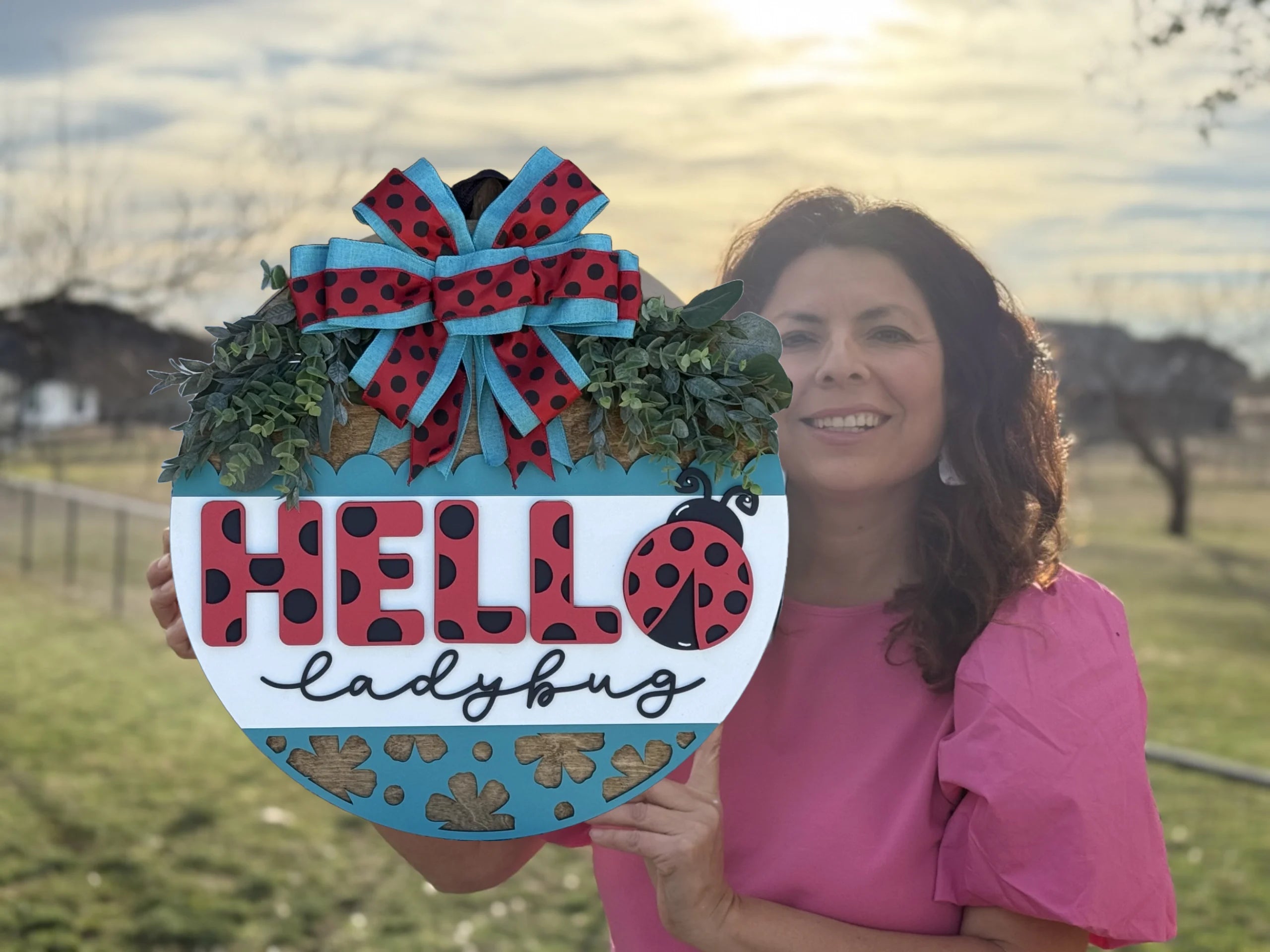 A woman in a pink blouse holds a decorative sign outdoors. The sign features HELLO ladybug with a red and black ladybug theme, and a bow with matching colors on top. Greenery surrounds the bow, and the sky is partly cloudy with the sun shining.