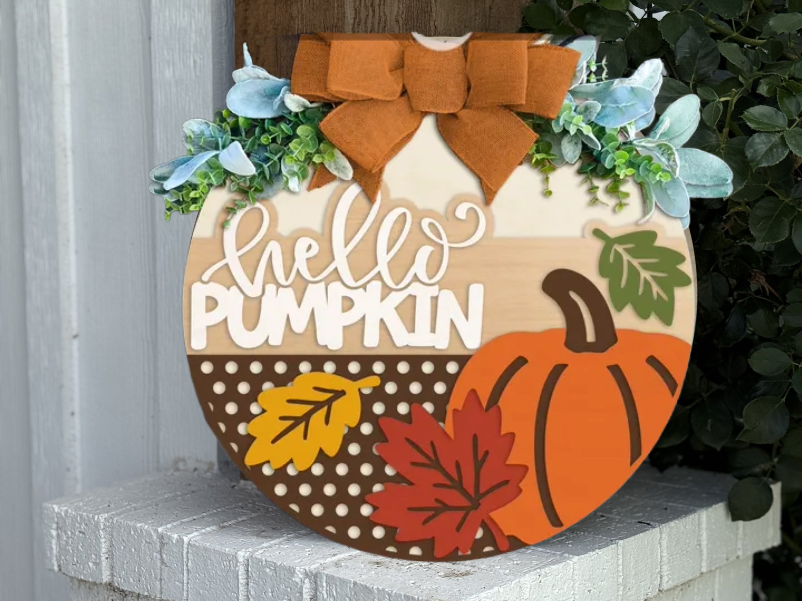 A round wooden sign decorated with a pumpkin, autumn leaves, greenery, a large orange bow, and the words hello PUMPKIN in white letters, displayed on a white brick ledge.