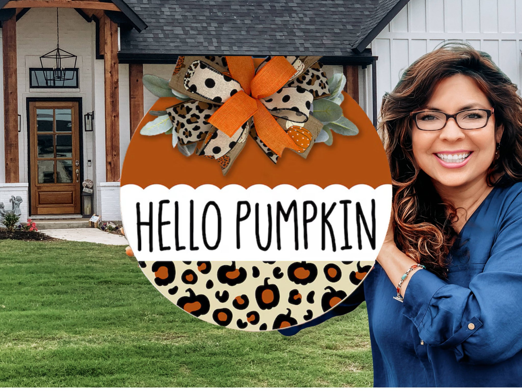 A smiling woman with wavy hair stands outside a modern house, holding a round sign decorated with leopard print that says HELLO PUMPKIN and has an orange and leopard print bow on top.
