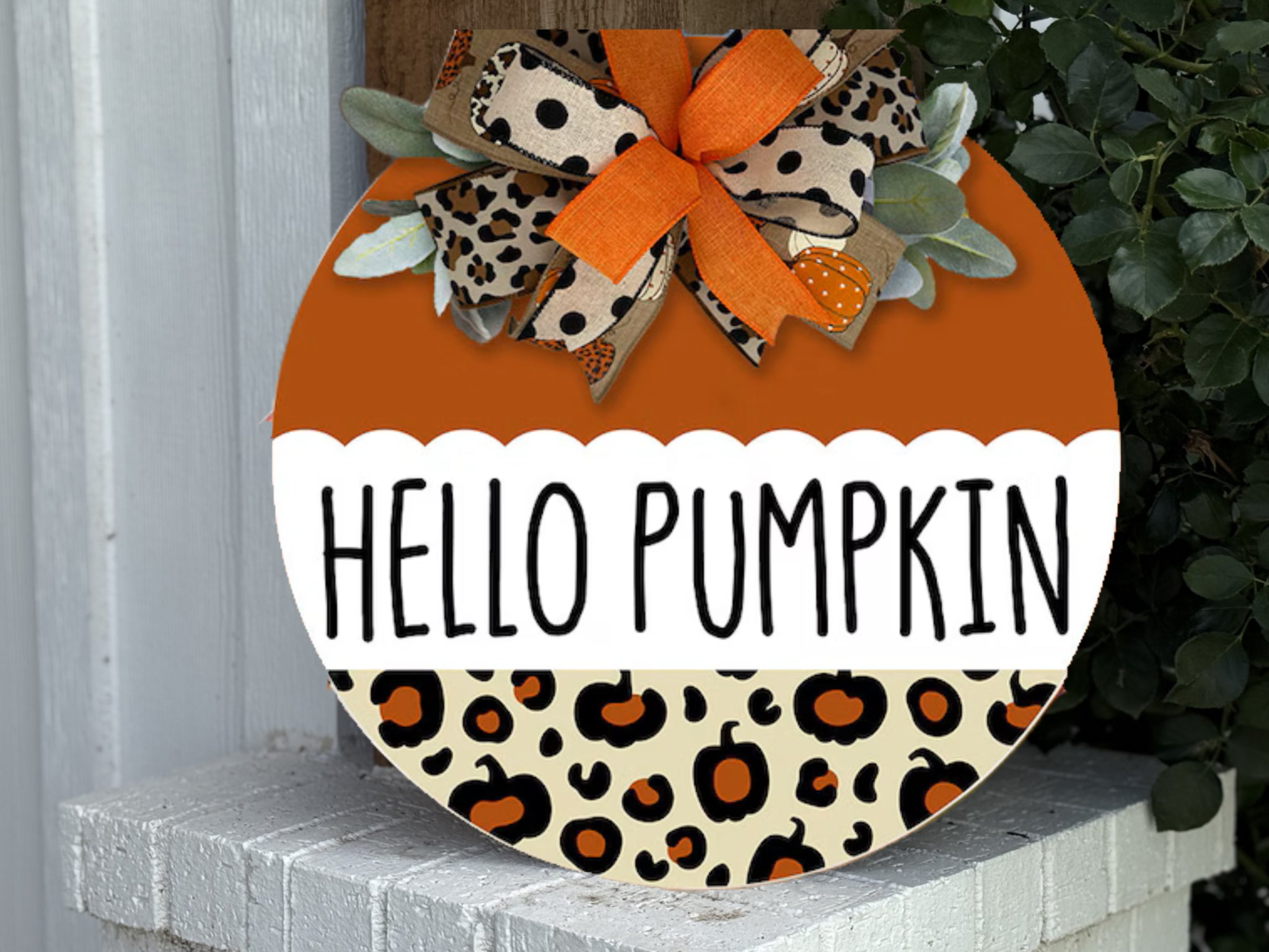 A round wooden sign with the words HELLO PUMPKIN in bold letters, decorated with a bow featuring orange and leopard print ribbons, and a base pattern of animal spots. Greenery peeks from behind the bow.