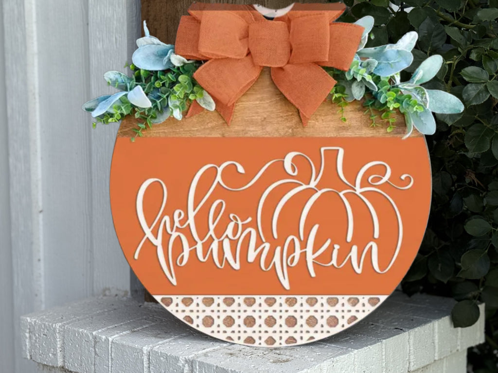 A round pumpkin-shaped wooden sign with “hello pumpkin” written in cursive, decorated with green leaves and a large orange bow at the top, leaning against a white brick surface.