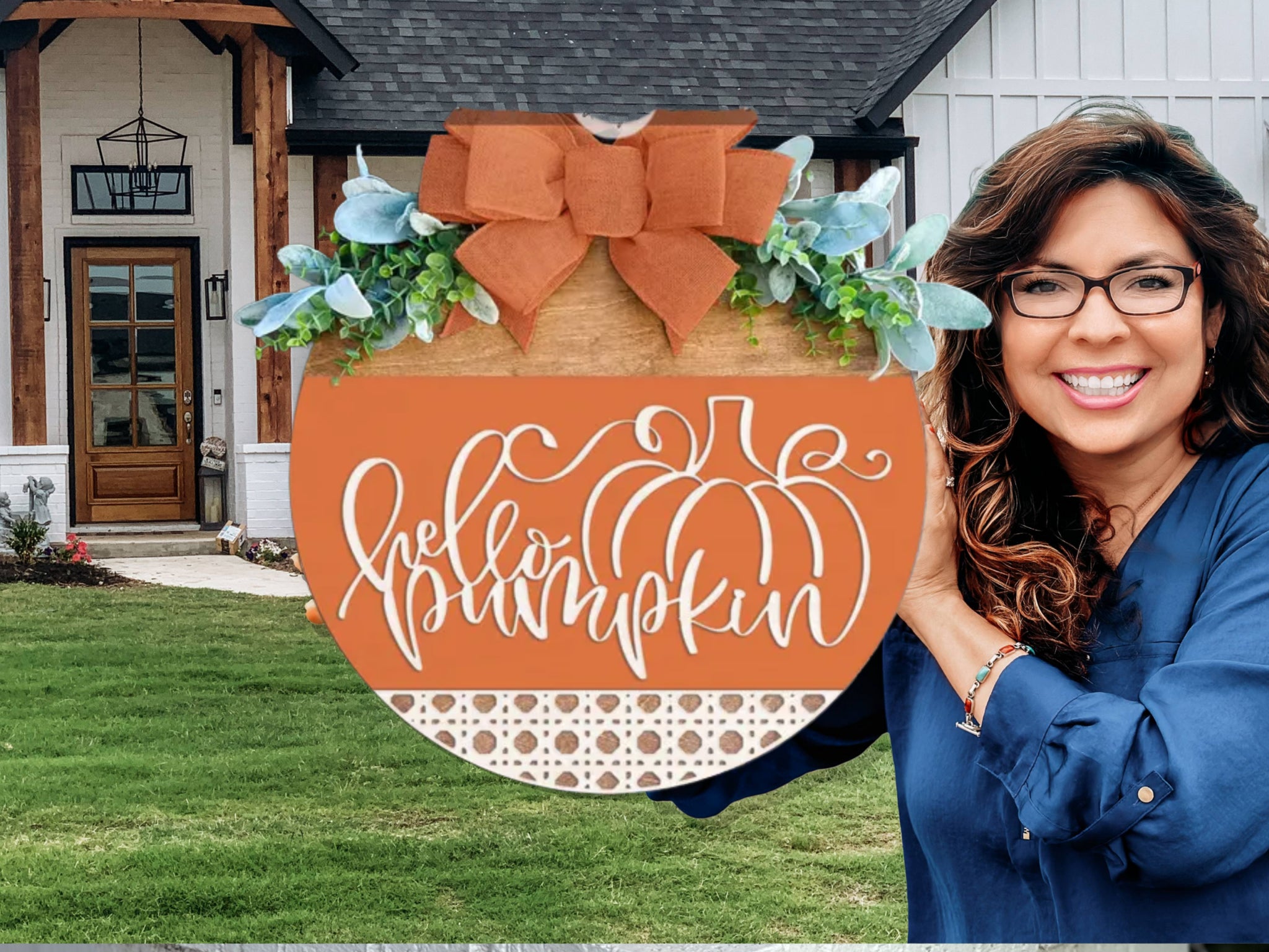 A smiling woman with glasses holds a round autumn sign reading hello pumpkin in front of a house with a green lawn. The sign features a pumpkin, greenery, and an orange bow.