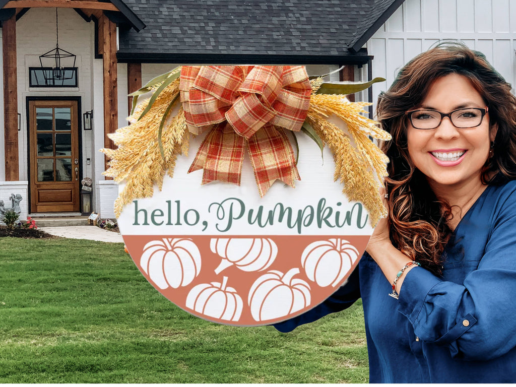 A smiling woman with glasses holds a round fall-themed sign that reads hello, Pumpkin with pumpkin illustrations and a plaid bow, standing outside a modern house with a green lawn.