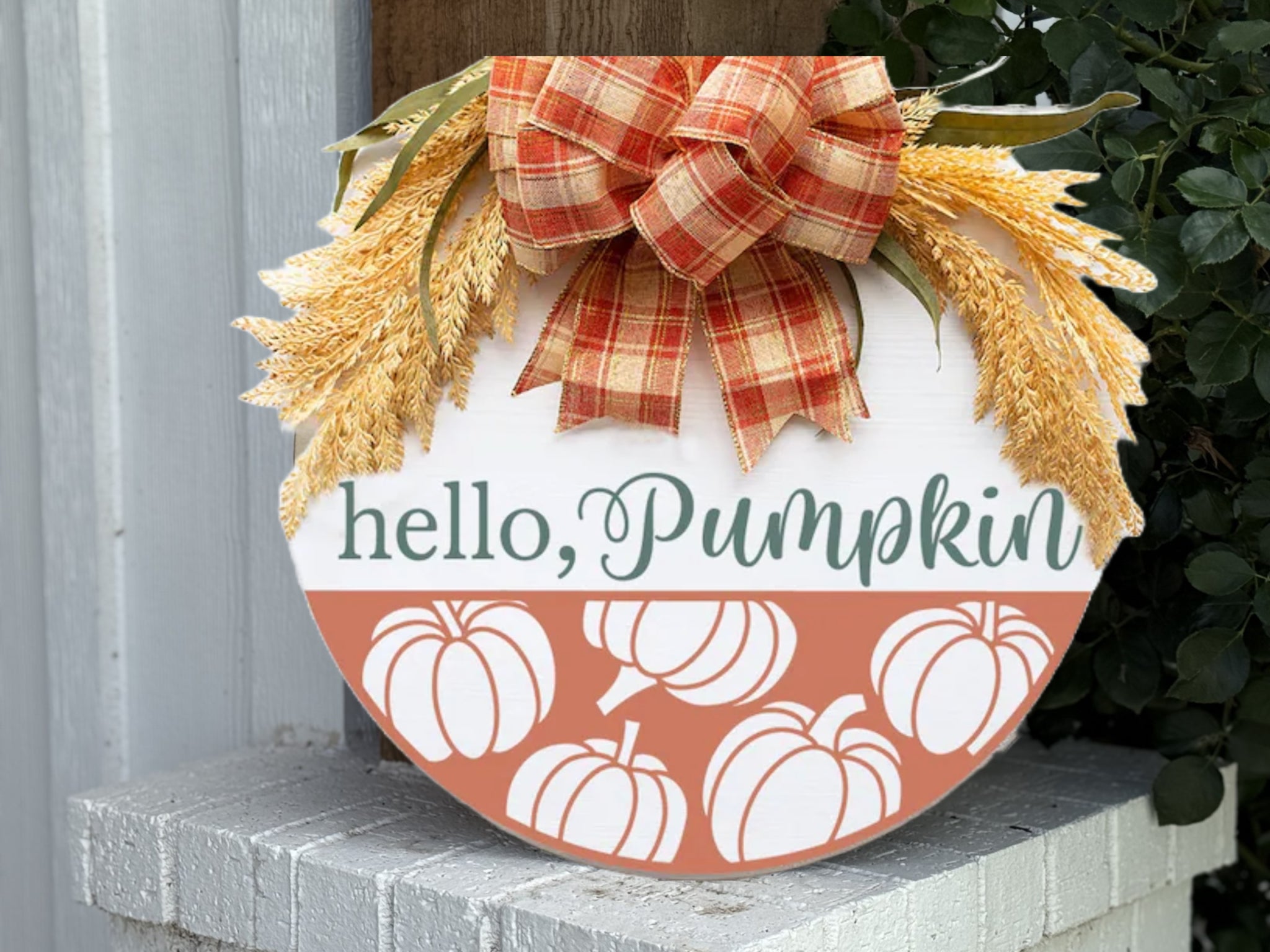 A round fall-themed sign decorated with a plaid bow, faux wheat, and leaves. The sign reads hello, Pumpkin and features white pumpkin illustrations on an orange background at the bottom.