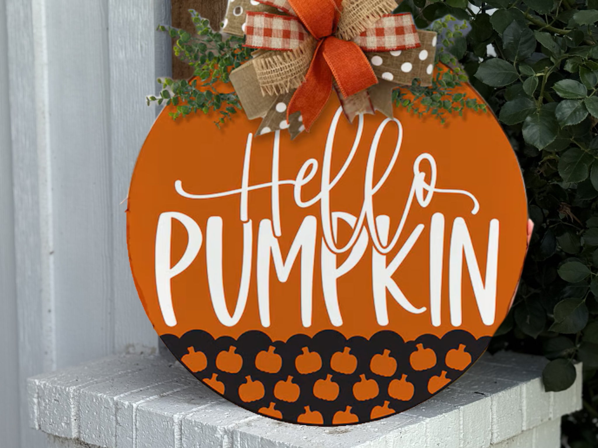 A round orange sign with the words Hello Pumpkin in white script, decorated with small pumpkin icons and topped with a bow made of burlap, red, and checkered ribbons, sits on a white brick surface near green plants.