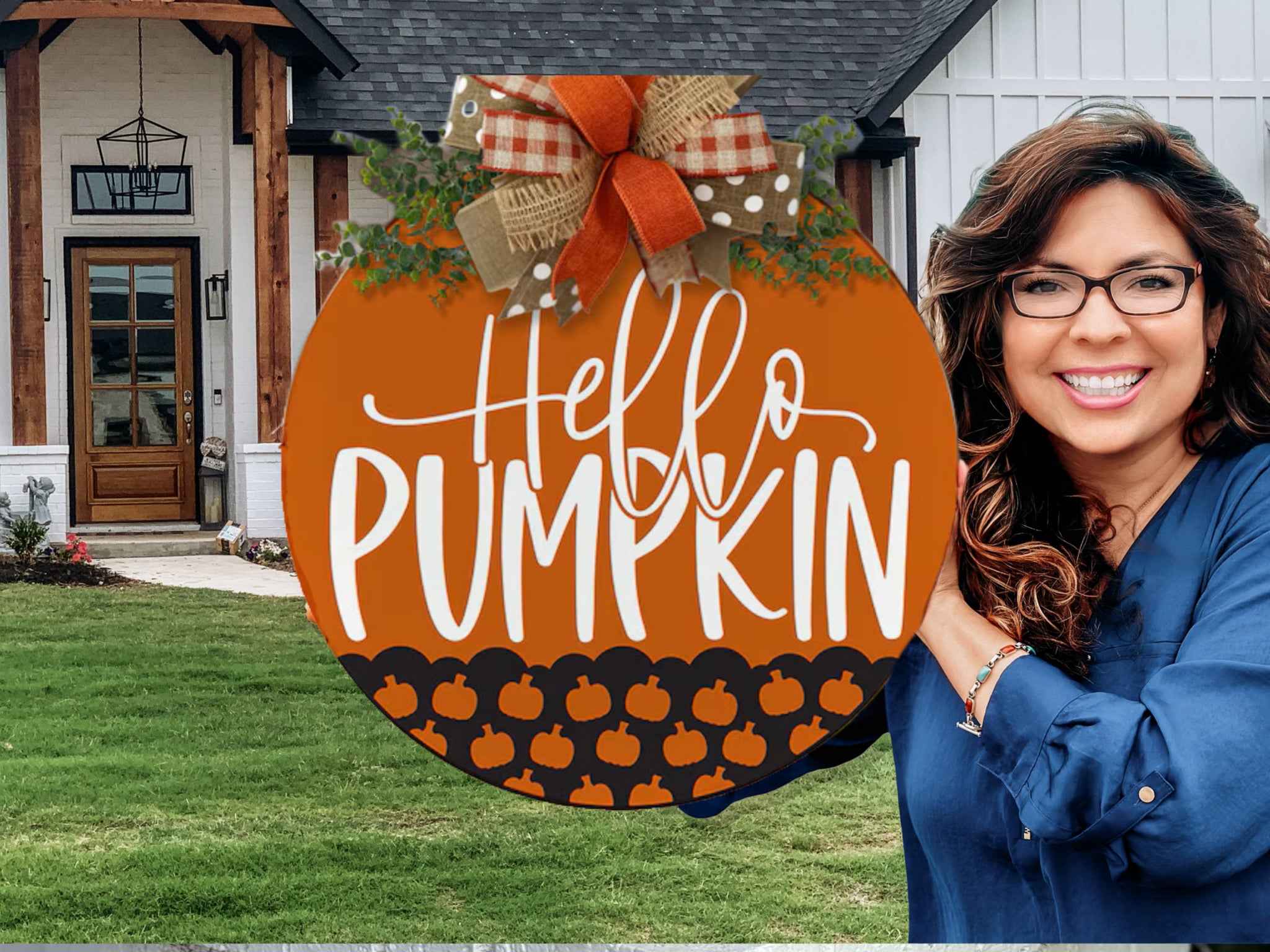 A woman with wavy brown hair and glasses smiles in front of a house, holding a round autumn sign that says Hello Pumpkin decorated with a bow and small pumpkins.