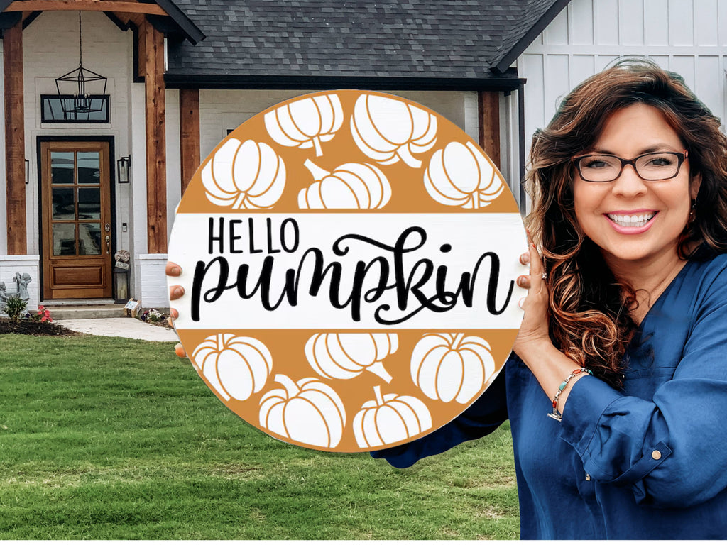 A smiling woman in glasses stands outside a house, holding a round sign decorated with white pumpkins and the words Hello Pumpkin in bold, playful lettering.