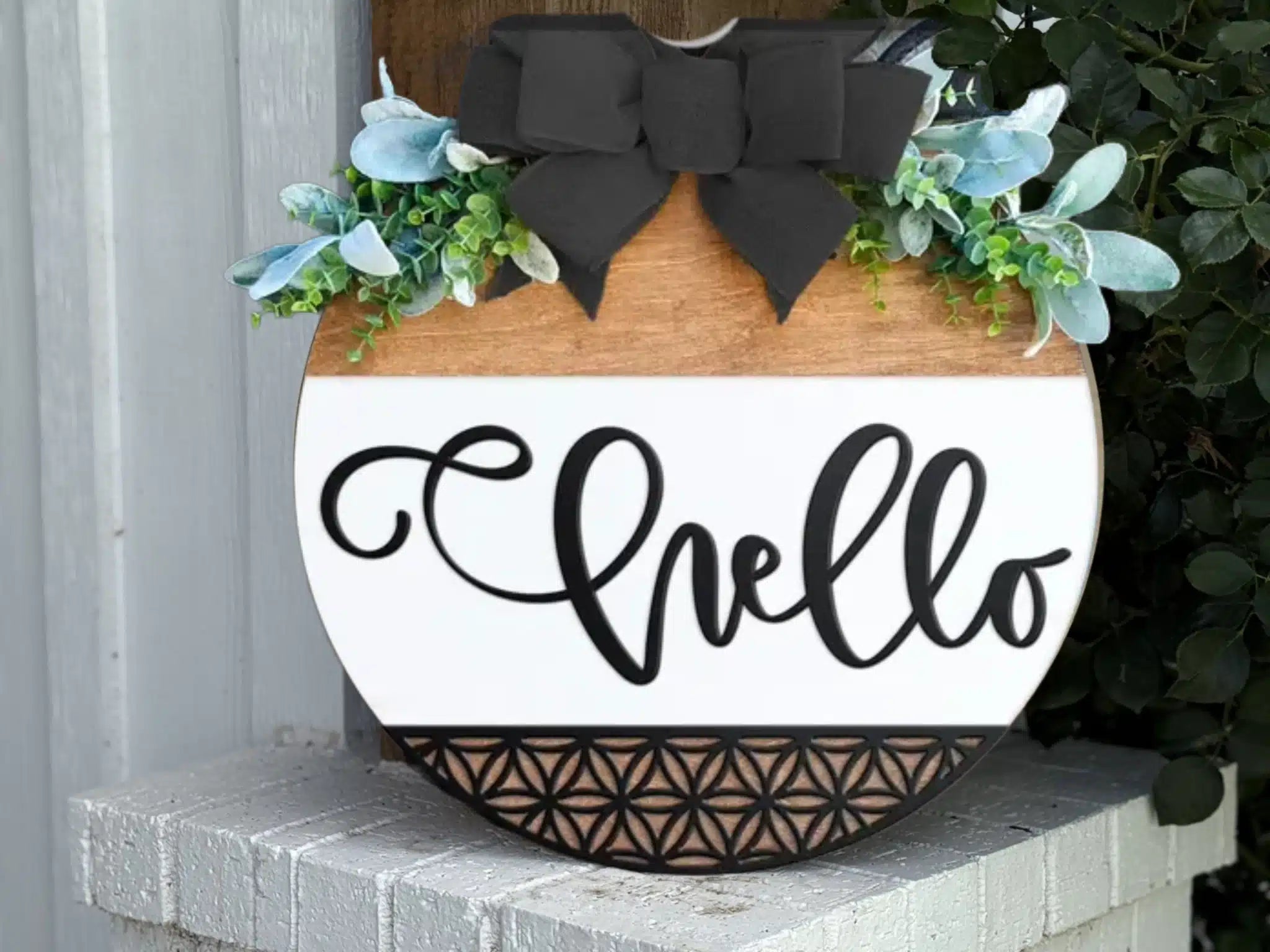 A round wooden sign with hello written in black script, decorated with a large black bow and green foliage at the top, and a geometric pattern at the bottom, is displayed on a white brick ledge.