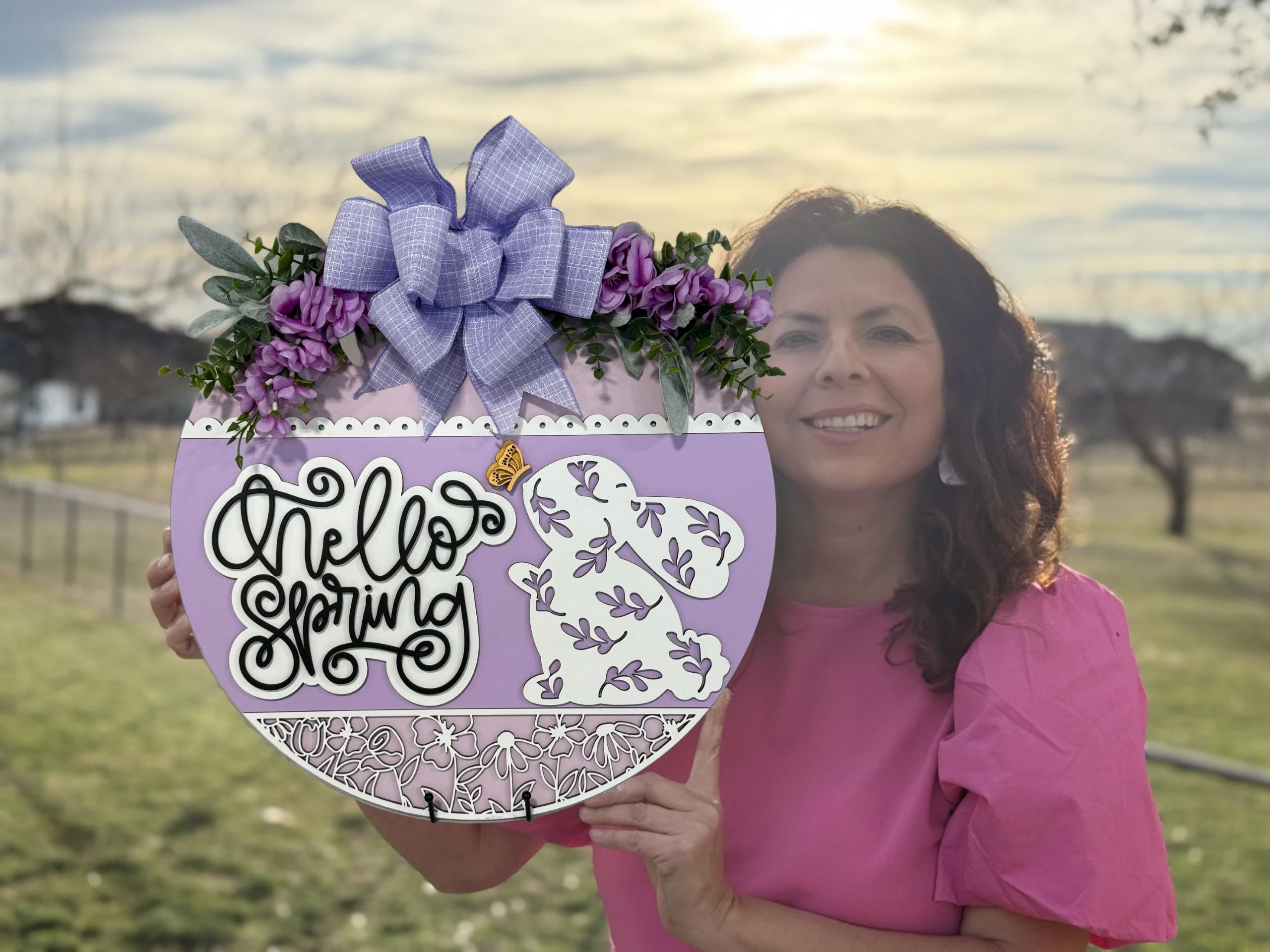 A woman in a pink shirt holds a decorative circular sign with Hello Spring written in cursive. The sign is adorned with purple flowers, a large purple bow, and has a bunny design. The sun is setting in the background over a grassy field and trees.