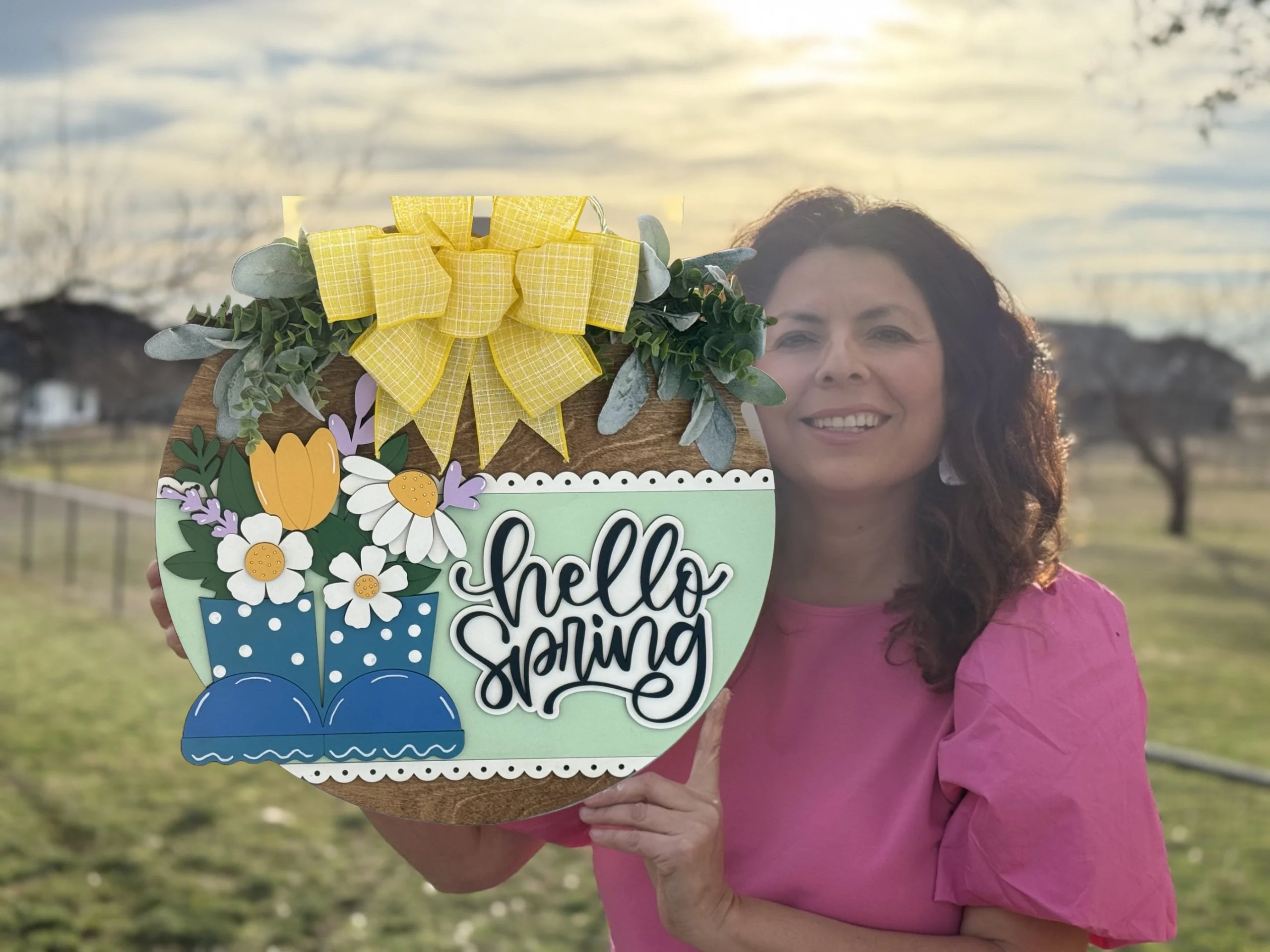 A woman outdoors holds a round spring-themed sign featuring yellow flowers, blue rain boots, and the words Hello Spring in cursive. The sign is adorned with a yellow checkered bow and greenery. She wears a pink blouse, and the background shows a sunlit field and cloudy sky.