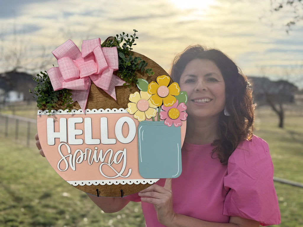 A woman in a pink blouse holds a round, decorative sign that reads Hello Spring. The sign features pink and yellow flowers, greenery, and a large pink bow. The background includes a grassy field and a bright, setting sun in a partly cloudy sky.