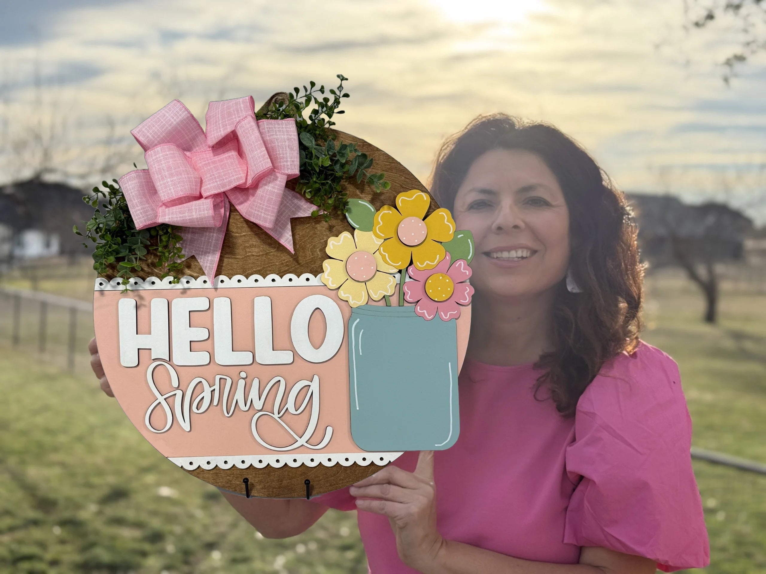A woman in a pink blouse holds a round, decorative sign that reads Hello Spring. The sign features pink and yellow flowers, greenery, and a large pink bow. The background includes a grassy field and a bright, setting sun in a partly cloudy sky.