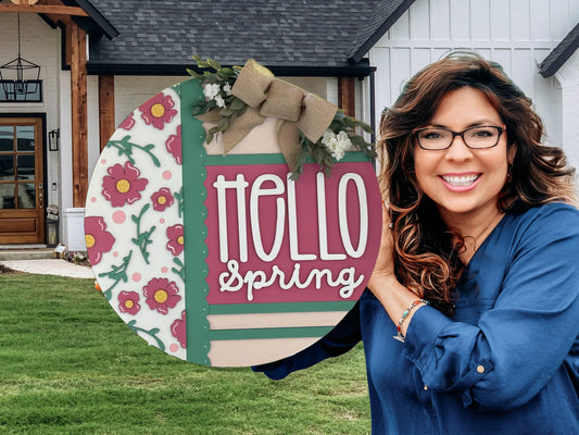 A smiling woman with long brown hair and glasses stands outside a house, holding a large round sign decorated with pink flowers, green stripes, and a bow. The sign reads Hello Spring in bold white and pink letters. The background shows a yard and porch.
