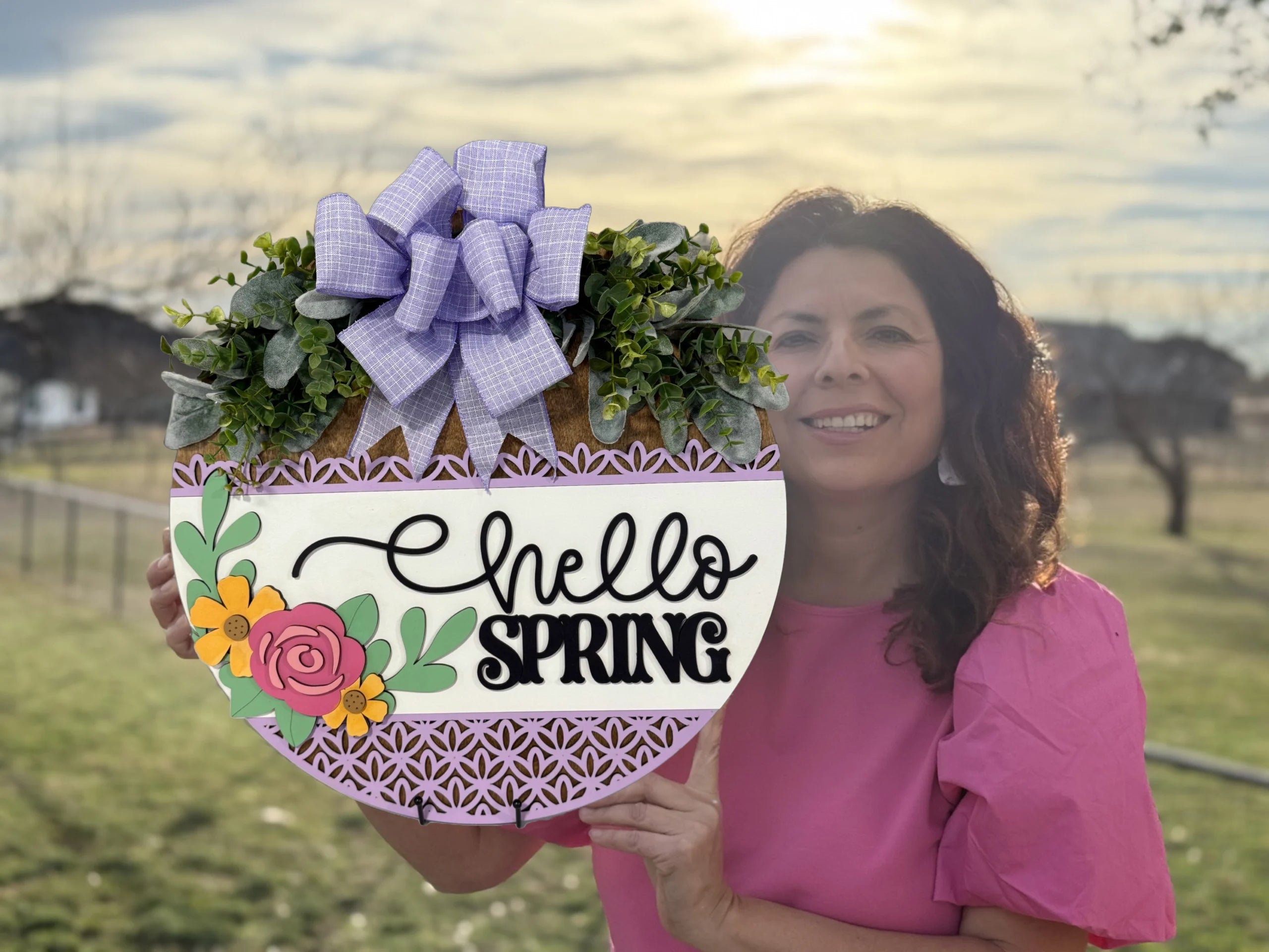 A woman in a pink shirt holds a decorative sign that says Hello Spring with colorful floral designs and green leaves. The sign features a checkered ribbon bow on top. She stands outdoors with a blurred background of a grassy field and trees under a partly cloudy sky.