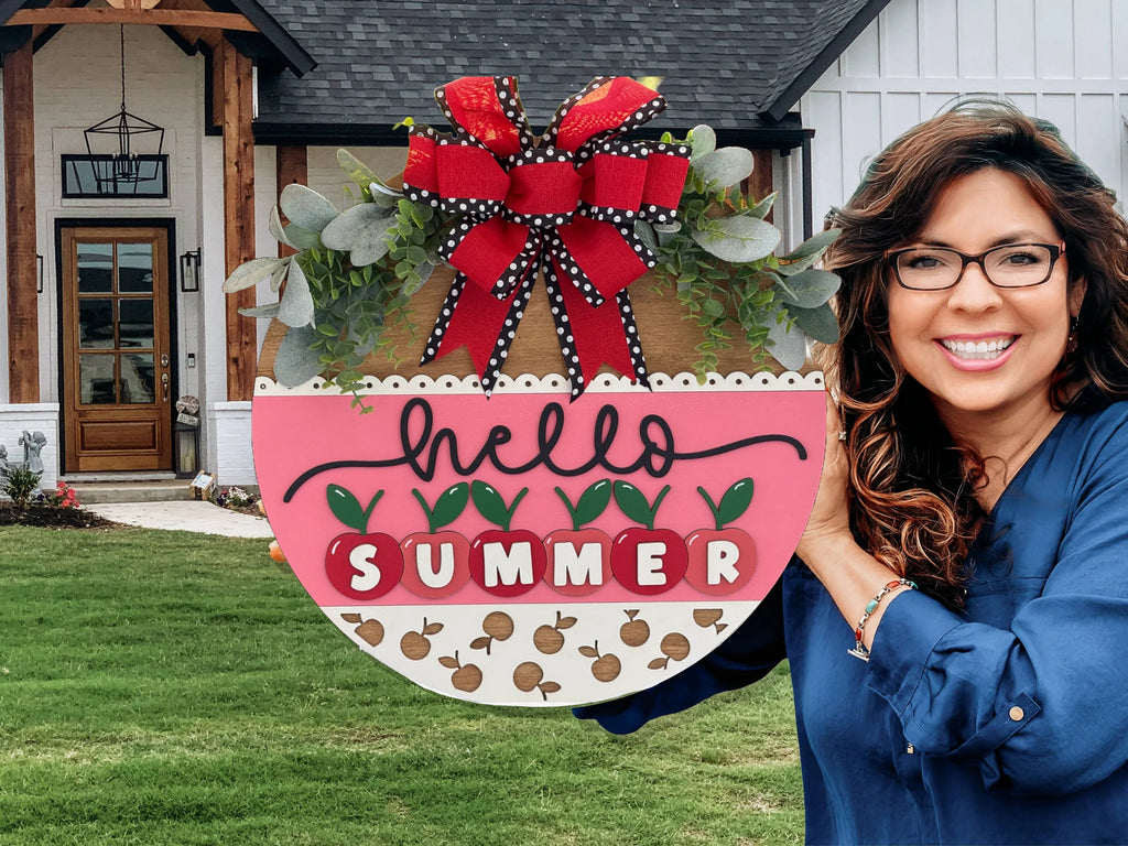A smiling woman with long brown hair and glasses holds a large round sign that reads “hello SUMMER” in pink, white, and red letters, decorated with greenery and a red and black bow, standing in front of a modern house with a manicured lawn.