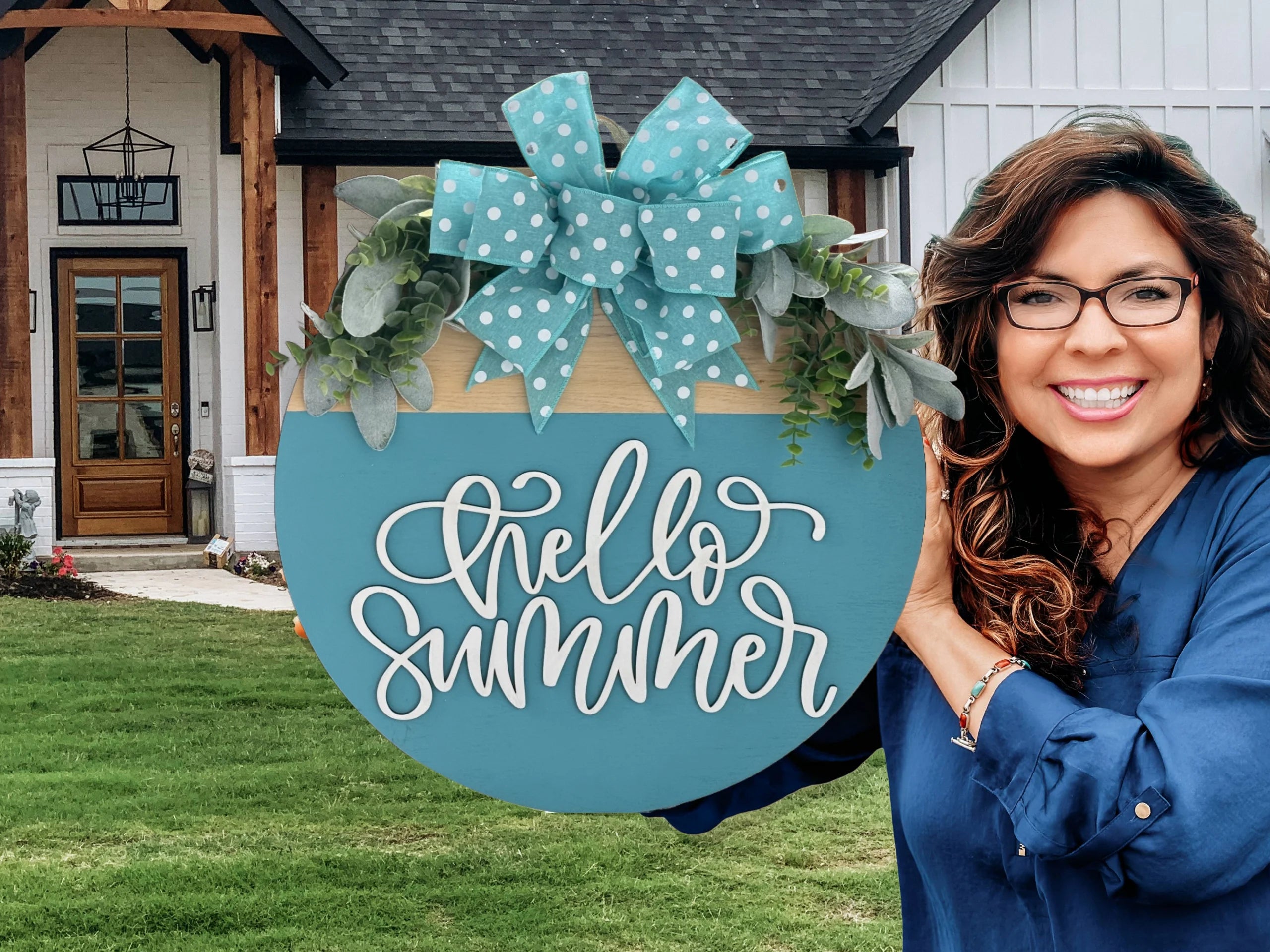 Woman smiling and holding a large blue sign with Hello Summer written on it in cursive. The sign is adorned with green foliage and a polka-dotted bow. She stands outside a modern house with a wooden entrance and well-manicured lawn. The background includes siding and a shingled roof.